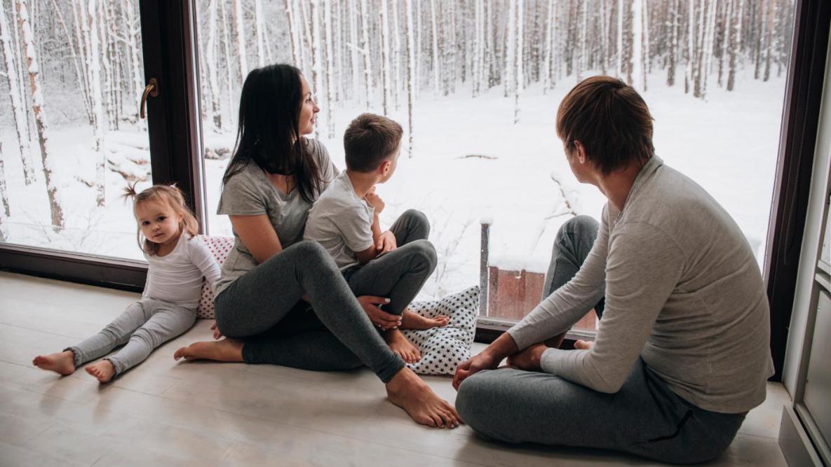 En familie på fire personer sitter på gulvet ved et stort vindu og kikker ut på vinterlandskap.