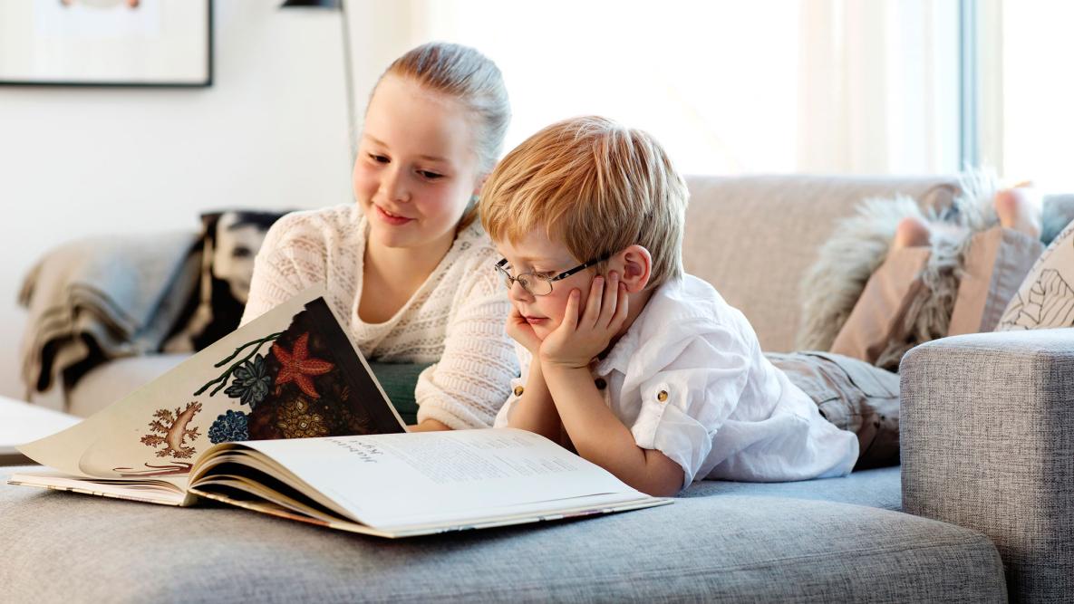 Miljøbilde av to små barn som leser i en bok i sofaen