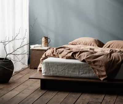 Seng med sengetøy i brun farge og en lys blålig farge på veggen. Lyse gardiner til venstre der solen skinner inn