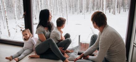 En familie på fire personer sitter på gulvet ved et stort vindu og kikker ut på vinterlandskap.